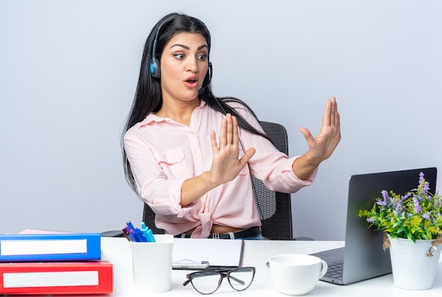 Bezpłatne zdjęcie młoda piękna kobieta w zwykłych ubraniach ze słuchawkami i mikrofonem siedzi przy stole z laptopem, patrząc na ekran, zmartwiona i zdezorientowana nad białą ścianą pracy w biurze
