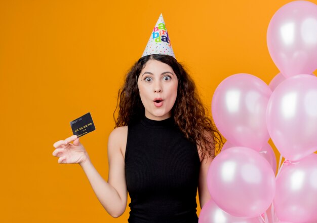 Młoda piękna kobieta z kręconymi włosami w świątecznej czapce, trzymając balony i wygląd karty kredytowej, zaskoczona koncepcja przyjęcia urodzinowego stojącego nad pomarańczową ścianą