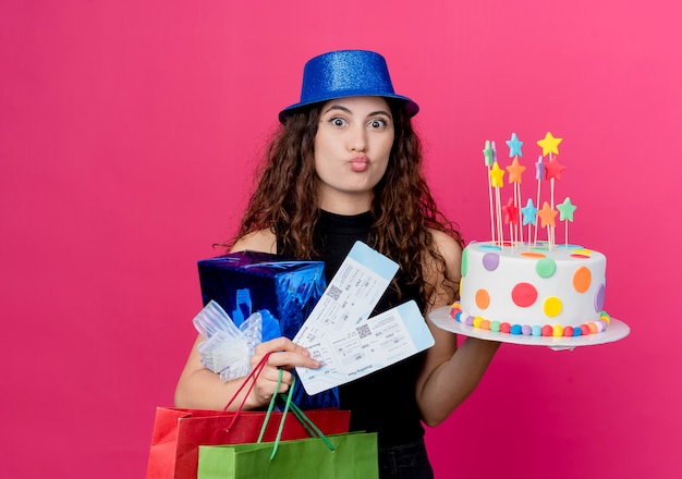 Młoda piękna kobieta z kręconymi włosami w świątecznym kapeluszu, trzymając pudełko na tort urodzinowy i bilety lotnicze koncepcja szczęśliwego i pozytywnego przyjęcia urodzinowego stojącego nad różową ścianą