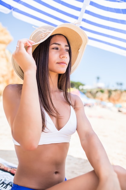 Młoda piękna Łacińska kobieta w bikini i słomkowy kapelusz siedzi pod parasolem na plaży w pobliżu wybrzeża morskiego.