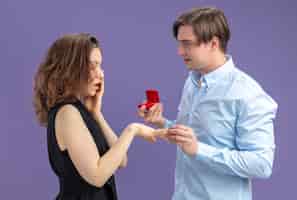 Bezpłatne zdjęcie młoda piękna para szczęśliwy mężczyzna składa propozycję z pierścionkiem zaręczynowym w czerwonym pudełku dla swojej uroczej zaskoczonej dziewczyny podczas walentynek stojących na niebieskim tle