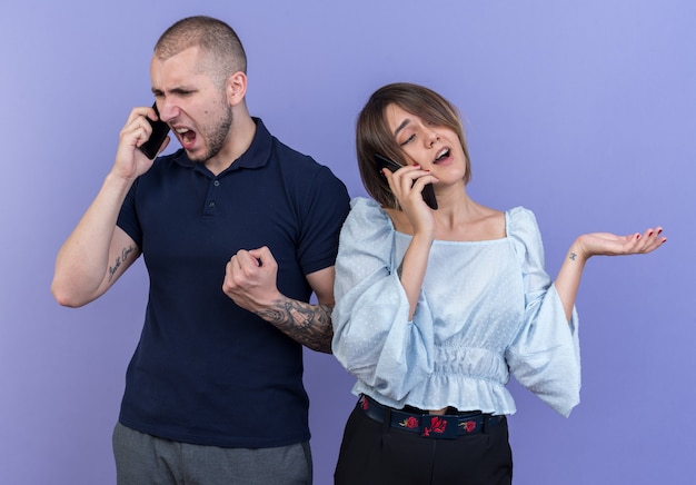 Bezpłatne zdjęcie młoda piękna para zły mężczyzna krzyczy podczas rozmowy przez telefon komórkowy, podczas gdy jego szczęśliwa dziewczyna uśmiecha się rozmawiając przez telefon komórkowy stojący nad niebieską ścianą
