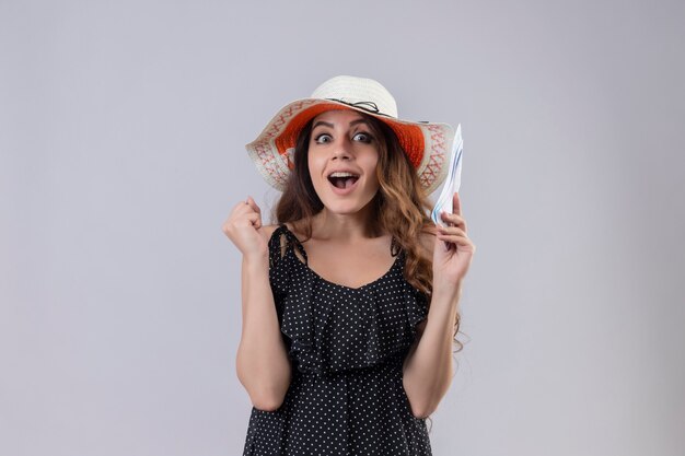 Młoda piękna podróżniczka w sukience w kropki w letnim kapeluszu trzymająca bilety lotnicze wyglądająca na podekscytowaną i szczęśliwą podnoszącą pięść, ciesząc się jej sukcesem i zwycięstwem stojącym na białym tle