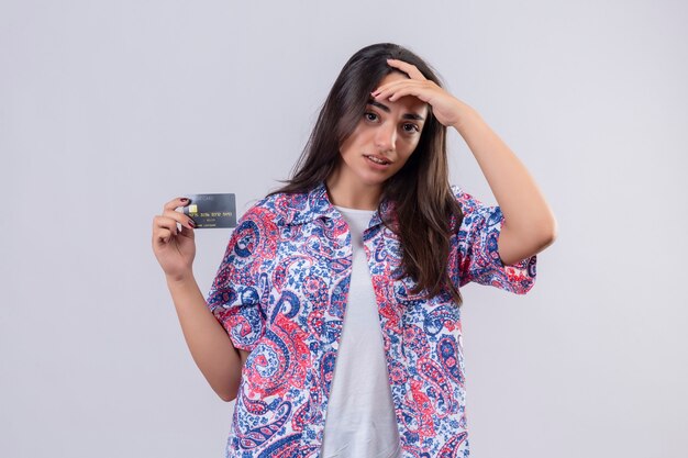 Młoda piękna podróżnik kobieta trzyma kartę kredytową, patrząc zdezorientowany dotykając głowy wątpliwe wyrażenie stojąc na białym tle