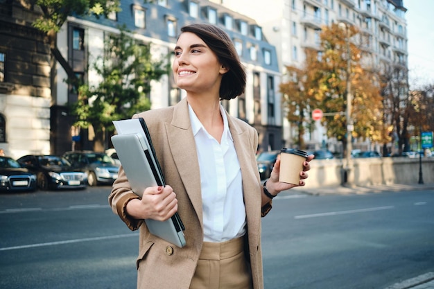 Młoda piękna szczęśliwa bizneswoman z laptopem i kawą, aby przejść na ulicę miasta