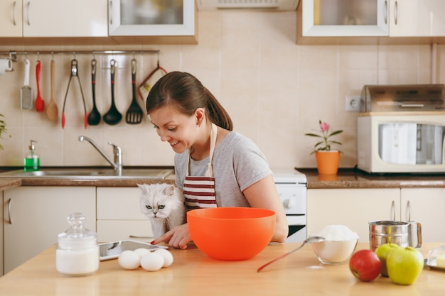 Młoda piękna szczęśliwa kobieta z białym kotem perskim szuka przepisu na ciastka na tablecie w kuchni. gotowanie w domu. przygotuj jedzenie.
