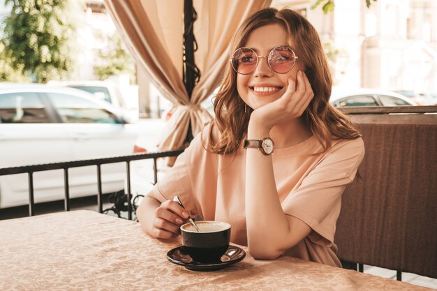 Młoda piękna uśmiechnięta hipster dziewczyna w modne letnie ubrania. Beztroski kobieta siedzi w kawiarni na tarasie werandy i pije kawę. Pozytywny model zabawy i marzeń