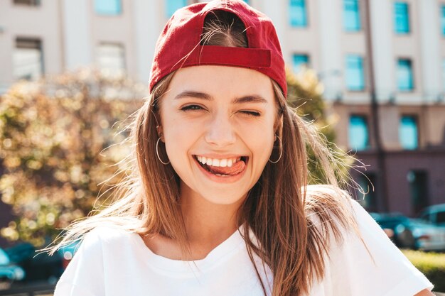 Młoda piękna uśmiechnięta kobieta hipster w modnej letniej białej koszulce Seksowna beztroska kobieta pozuje na tle ulicy w czapce o zachodzie słońca Pozytywny model na zewnątrz Wesoły i szczęśliwy Winking