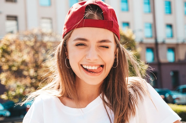 Młoda piękna uśmiechnięta kobieta hipster w modnej letniej białej koszulce Seksowna beztroska kobieta pozuje na tle ulicy w czapce o zachodzie słońca Pozytywny model na zewnątrz Wesoły i szczęśliwy Winking