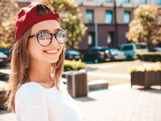 Bezpłatne zdjęcie młoda piękna uśmiechnięta kobieta hipster w modnej letniej białej koszulce seksowna beztroska kobieta pozuje na tle ulicy w czapce o zachodzie słońca pozytywny model na zewnątrz wesoły i szczęśliwy