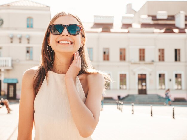 Młoda piękna uśmiechnięta kobieta hipster w modnych letnich ubraniach Seksowna beztroska kobieta pozuje na tle ulicy o zachodzie słońca Pozytywny model na zewnątrz Wesoły i szczęśliwy w okularach przeciwsłonecznych