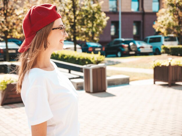 Bezpłatne zdjęcie młoda piękna uśmiechnięta kobieta hipster w modnych letnich ubraniach seksowna beztroska kobieta pozuje na tle ulicy w czapce o zachodzie słońca pozytywny model na zewnątrz wesoły i szczęśliwy w okularach przeciwsłonecznych