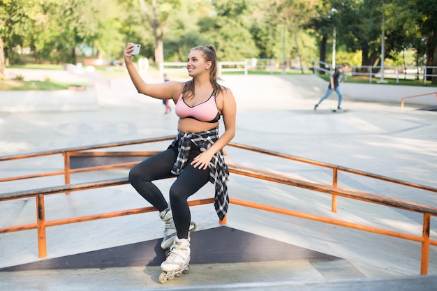 Młoda piękna uśmiechnięta kobieta plus size w różowych sportowych topleggingach i wrotkach opierając się na poręczy szczęśliwie robi zdjęcie na telefonie komórkowym podczas spędzania czasu w skate parku