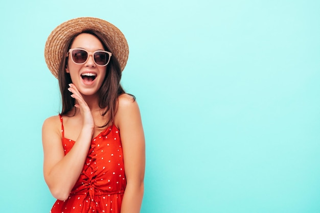 Młoda piękna uśmiechnięta kobieta w modnej letniej czerwonej sukience Seksowna beztroska kobieta pozuje w pobliżu niebieskiej ściany w studio Pozytywna modelka brunetka bawi się w kapeluszu i okularach przeciwsłonecznych Krzyki i krzyki