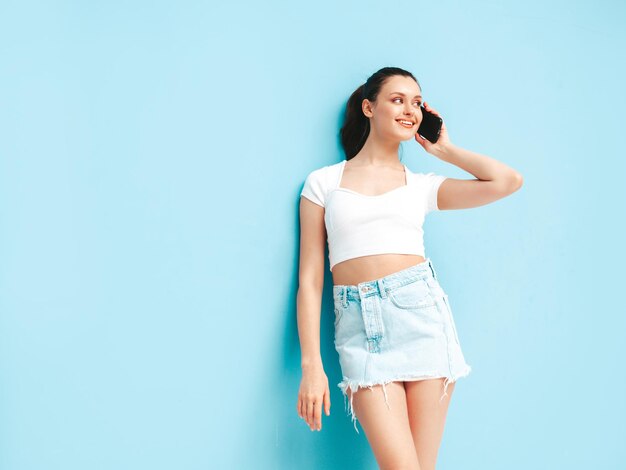 Młoda piękna uśmiechnięta kobieta w modnej letniej spódnicy jeansowej i topowych ubraniach Seksowna beztroska kobieta pozuje w pobliżu niebieskiej ściany w studio Pozytywna brunetka modelka bawi się rozmawiając na smartfonie