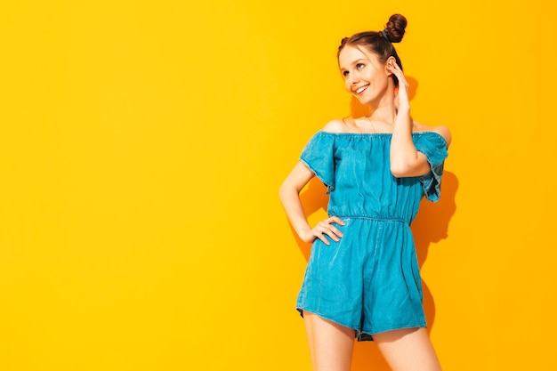 Młoda piękna uśmiechnięta kobieta w modnych letnich dżinsach kombinezonach Seksowna beztroska kobieta z dwoma rogami fryzura pozowanie w pobliżu żółtej ściany w studio Pozytywny model zabawy na białym tle