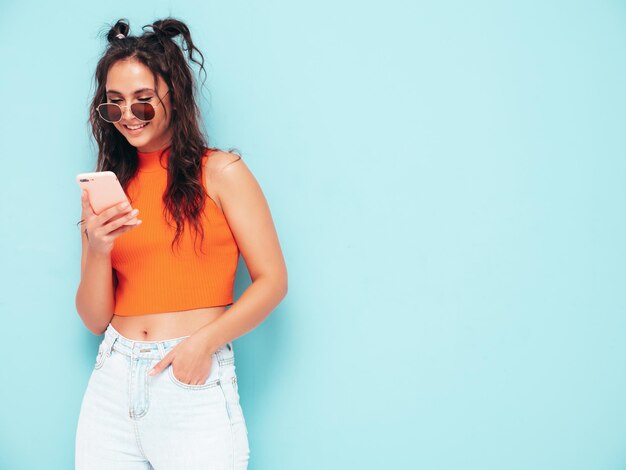 Młoda piękna uśmiechnięta kobieta w modnych letnich pomarańczowych ubraniach i dżinsach Seksowna beztroska kobieta pozuje w pobliżu niebieskiej ściany w studio Patrząc na ekran smartfona