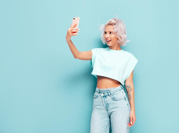 Młoda piękna uśmiechnięta kobieta w modnych letnich ubraniach Seksowna beztroska kobieta pozuje w pobliżu niebieskiej ściany w studio Pozytywna blond modelka bawi się i wariuje Wesoła i szczęśliwa Robienie zdjęć selfie
