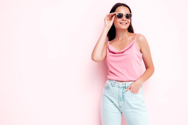 Młoda piękna uśmiechnięta kobieta w modnych letnich ubraniach Seksowna beztroska kobieta pozuje w pobliżu różowej ściany w studio Pozytywna brunetka modelka bawi się i wariuje Wesoła i szczęśliwa w okularach przeciwsłonecznych