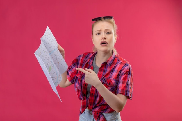 Młoda Podróżniczka W Czerwonej Koszuli I Okularach Na Głowie Wskazuje Na Mapę Na Odosobnionej Różowej ścianie