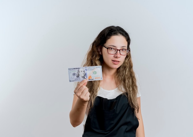 Młoda profesjonalna fryzjerka kobieta w fartuch pokazuje pieniądze patrząc z przodu z poważną twarzą stojącą na białej ścianie