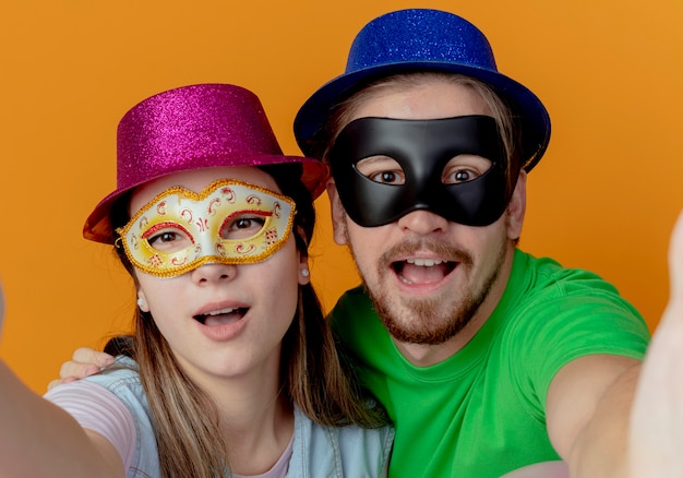 Młoda radosna para w różowych i niebieskich czapkach zakłada maskaradowe maski na oczy udaje, że trzyma i wygląda na odizolowaną na pomarańczowej ścianie