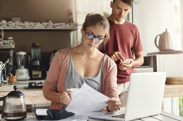 Młoda rodzina w obliczu kłopotów finansowych. Kobieta w okularach czytająca informacje na kartce papieru ze skupionym spojrzeniem, płacąca rachunki za media przez internet przy użyciu laptopa i kalkulatora