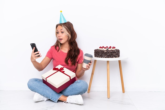 Młoda rosjanka świętująca urodziny, siedząca na podłodze na białym tle, trzymająca kawę na wynos i telefon komórkowy