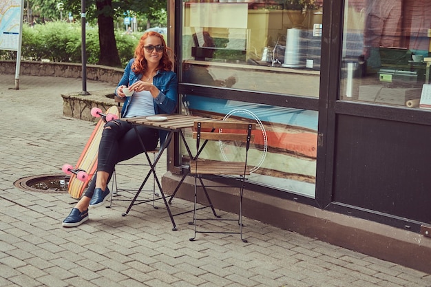 Bezpłatne zdjęcie młoda ruda dziewczyna pije kawę, siedząc w pobliżu kawiarni, relaksując się po jeździe na deskorolce.