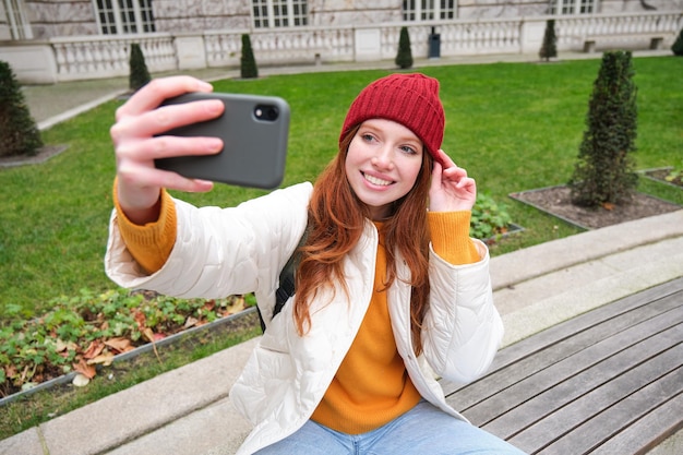 Bezpłatne zdjęcie młoda rudowłosa dziewczyna siedzi na ławce w parku i robi selfie, robiąc sobie zdjęcie z smartph