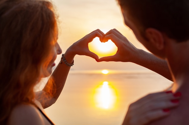 Młoda seksowna romantyczna para zakochanych szczęśliwy na letniej plaży razem dobrze się bawić na sobie kostiumy kąpielowe pokazując znak serca na zachodzie słońca
