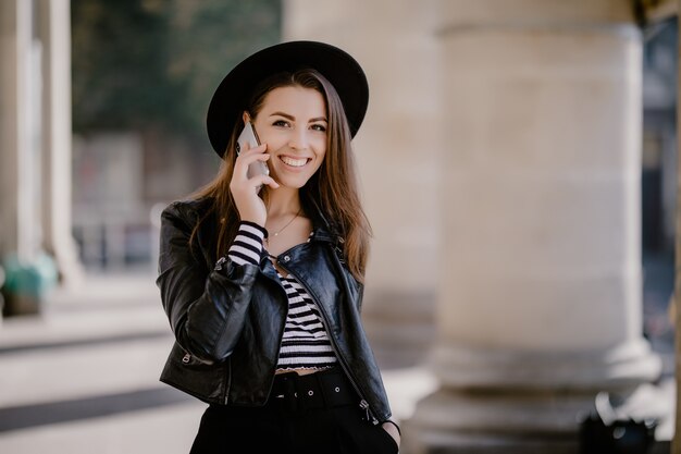 Młoda śliczna brązowowłosa dziewczyna w skórzanej kurtce, czarnym kapeluszu na miejskiej promenadzie prowadzi rozmowę telefoniczną