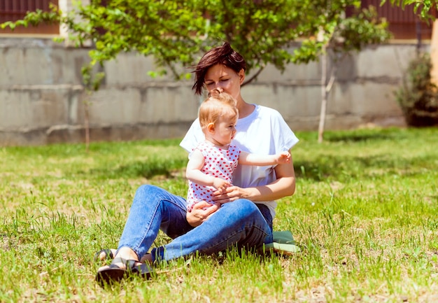 Młoda słodka mama siedzi na trawie w ogrodzie, trzymając córkę i coś do niej mówiąc. śliczna dziewczyna w letniej sukience w ramionach matki. letni czas rodzinny i koncepcja świeżego powietrza