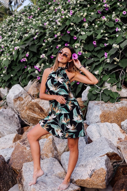 Młoda spokojna wytatuowana kobieta w letniej tropikalnej krótkiej sukience na kamienistej plaży z zielonym krzewem i fioletowymi różowymi kwiatami