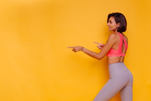 Młoda, sprawna, dość silna kobieta ubrana w sportowe stroje, stylowy top i legginsy, pozuje przed żółtą ścianą