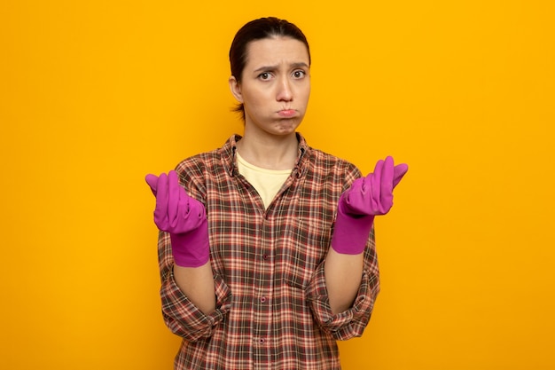 Młoda sprzątaczka w zwykłych ubraniach w gumowych rękawiczkach niezadowolona dmuchanie w policzki ocierające się o palce stojące nad pomarańczową ścianą