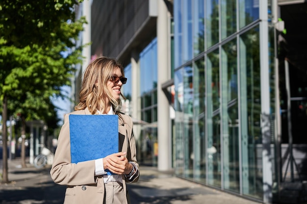 Bezpłatne zdjęcie młoda sprzedawczyni idzie do pracy w okularach przeciwsłonecznych i garniturze, trzymając folder z dokumentami chodzącymi po ulicy