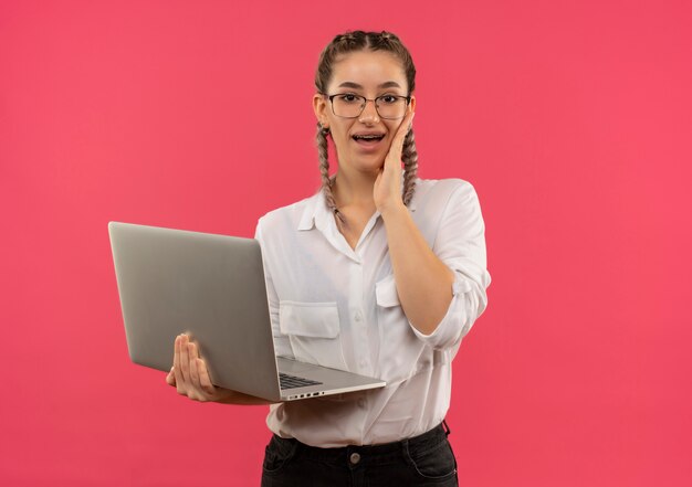 Młoda studentka dziewczyna w okularach z warkoczykami w białej koszuli trzymając laptopa patrząc do przodu zaskoczona i szczęśliwa stojąc nad różową ścianą