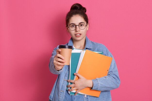 Młoda studentka o ciemnych włosach oferuje kawę na wynos, trzymając w rękach papierową teczkę