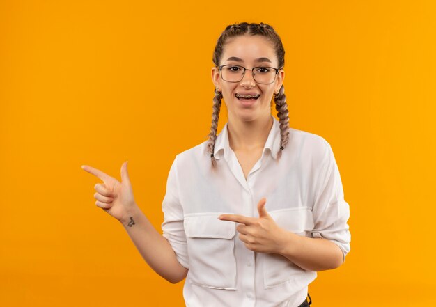 Młoda studentka w okularach z warkoczykami w białej koszuli uśmiechnięta, patrząc do przodu, wskazująca palcami wskazującymi na bok, stojąc nad pomarańczową ścianą