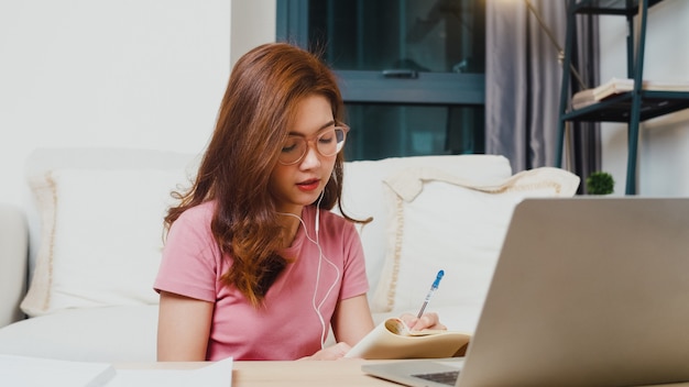 Młoda studentka z Azji nosi słuchawki do nauki na odległość z nauczycielem online na laptopie w salonie w domu w nocy. Dystans społeczny, kwarantanna w celu zapobiegania koronawirusom.