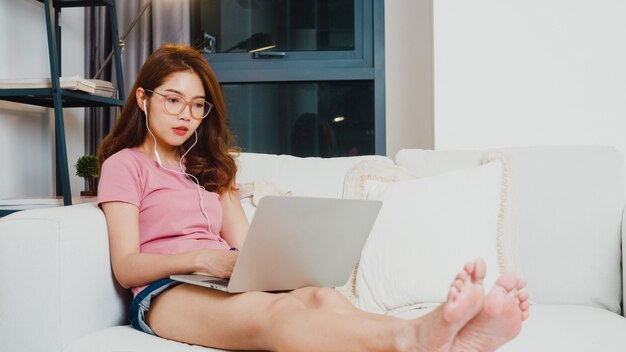 Młoda studentka z Azji nosi słuchawki do nauki na odległość z nauczycielem online na laptopie w salonie w domu w nocy. Dystans społeczny, kwarantanna w celu zapobiegania koronawirusom.