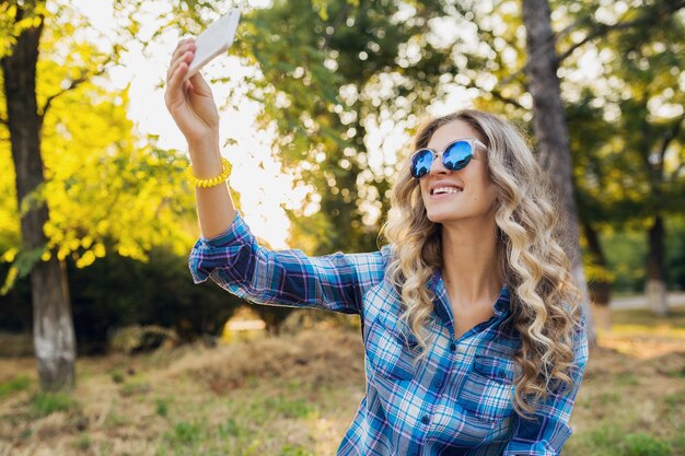 Młoda stylowa atrakcyjna uśmiechnięta blond kobieta siedzi w parku co selfie zdjęcie na telefon