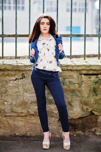 Młoda stylowa brunetka dziewczyna na koszuli spodnie jeansowa kurtka i wysokie obcasy buty pozowane tło żelazny płot Koncepcja modelu mody ulicznej