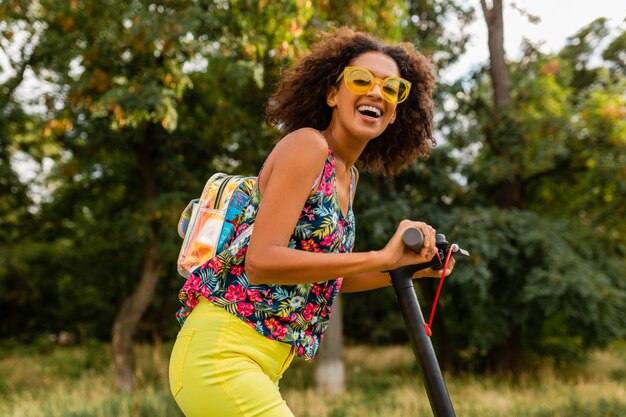 Młoda stylowa czarna kobieta bawi się w parku, jeżdżąc na hulajnodze elektrycznej w letnim stylu mody, kolorowy strój hipster, ubrany w plecak i żółte okulary przeciwsłoneczne