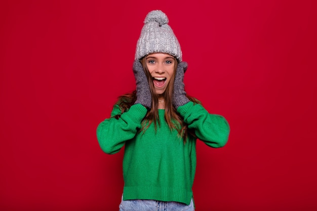 Młoda stylowa dziewczyna ubrana w szarą czapkę zimową i zielony sweter, pozowanie na białym tle z zaskoczonymi prawdziwymi emocjami. Obraz zabawne kobiety, zabawy z potrząsaniem włosów