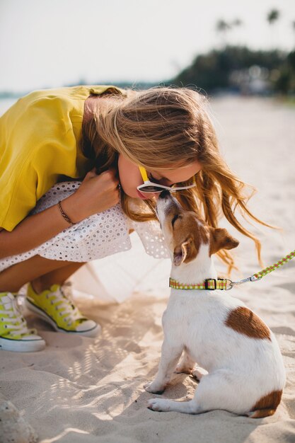 Młoda stylowa hipster kobieta trzyma chodzenie grając psa szczeniaka jack russell, tropikalny park, uśmiechnięty i baw się, wakacje, okulary przeciwsłoneczne, czapka, żółta koszula, piasek na plaży