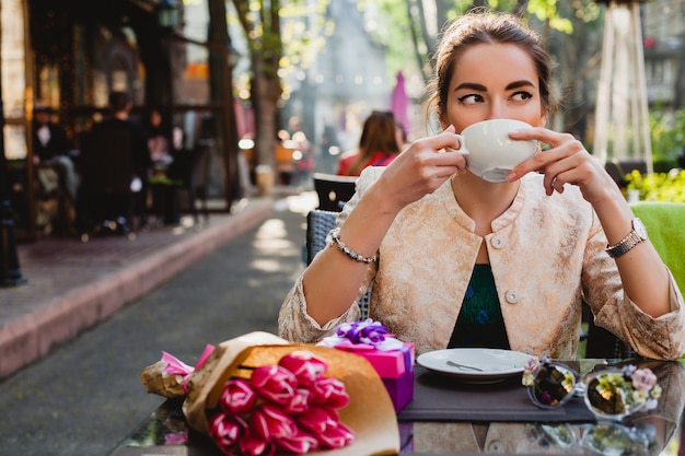 Młoda stylowa kobieta siedzi w kawiarni, trzymając filiżankę cappuccino