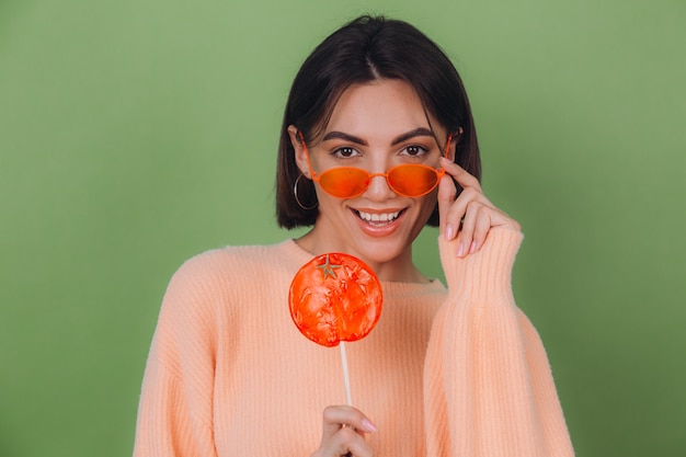 Młoda stylowa kobieta w dorywczo brzoskwiniowy sweter i pomarańczowe okulary na białym tle na zielonej oliwkowej ścianie z pomarańczowym lizakiem pozytywny uśmiech miejsca na kopię
