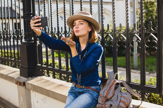 Młoda stylowa ładna kobieta wysyłająca buziaka przez telefon, ubrana w dżinsową koszulę i dżinsy